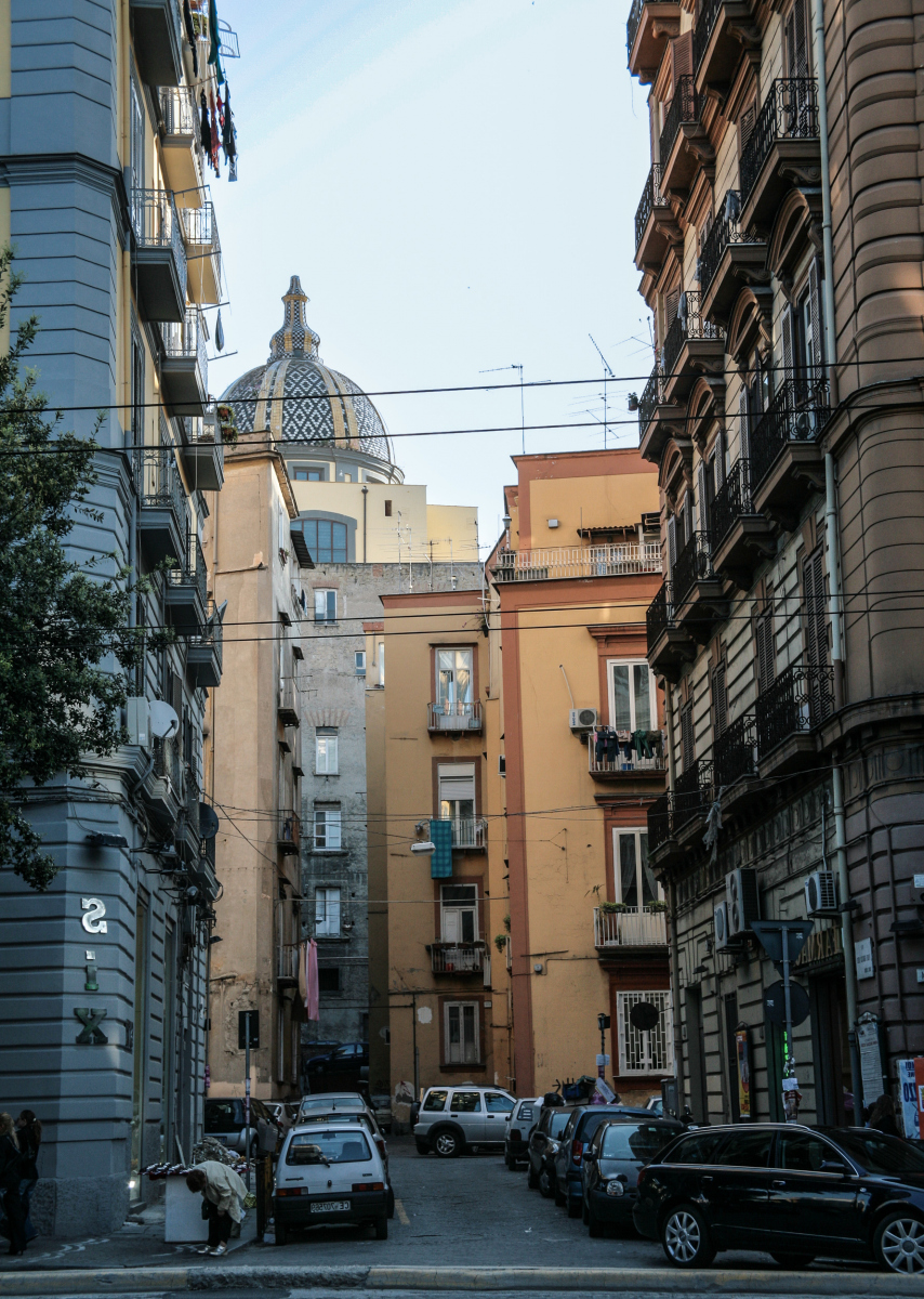 eine Gasse in Neapel, die direkt auf die markante Kuppel der Kirche Santi Marcellino e Festo blickt. Das Bild ist charakteristisch für das historische Zentrum von Neapel, einem UNESCO-Weltkulturerbe: Die Aufnahme entstand in der Via Leopoldo RodinòKirche Santi Marcellino e Festo: Die Kirche und das dazugehörige Kloster sind bedeutende barocke Bauwerke in Neapel. Der Komplex stammt ursprünglich aus dem 10. Jahrhundert und wurde im Laufe der Zeit mehrfach umgebaut und erweitert.Heutige Nutzung: Große Teile werden heute von der Universität Neapel Federico II genutzt