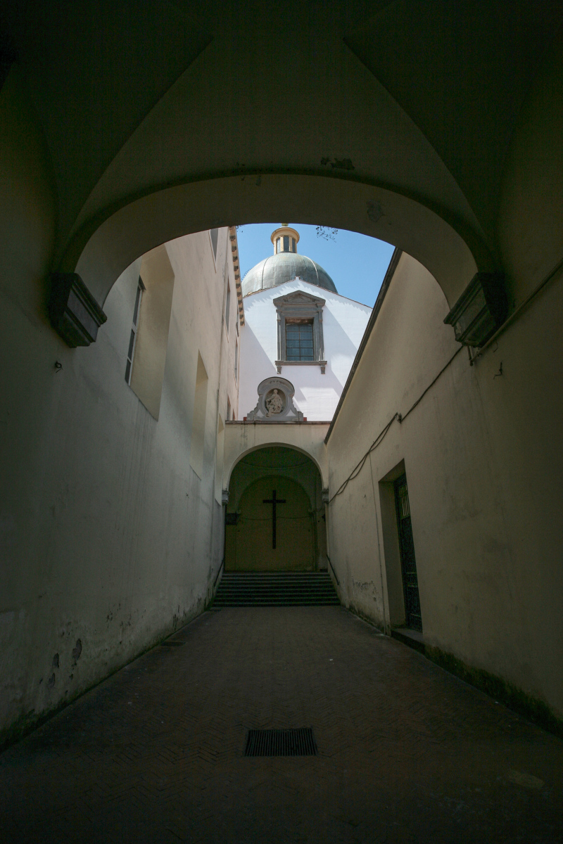 Das Bild zeigt den Durchgang zur Chiesa dei Girola. Blick durch einen gewölbten Gang auf die Kuppel der Kirche. Die Kirche und das angrenzende Kloster befinden sich an der Piazza Girolamini im historischen Zentrum in der Nähe des Doms. Die Kirche ist ein bedeutendes Beispiel der neapolitanischen Barockarchitektur. Der gesamte Komplex, der im späten 16. und frühen 17. Jahrhundert erbaut wurde, ist monumental.