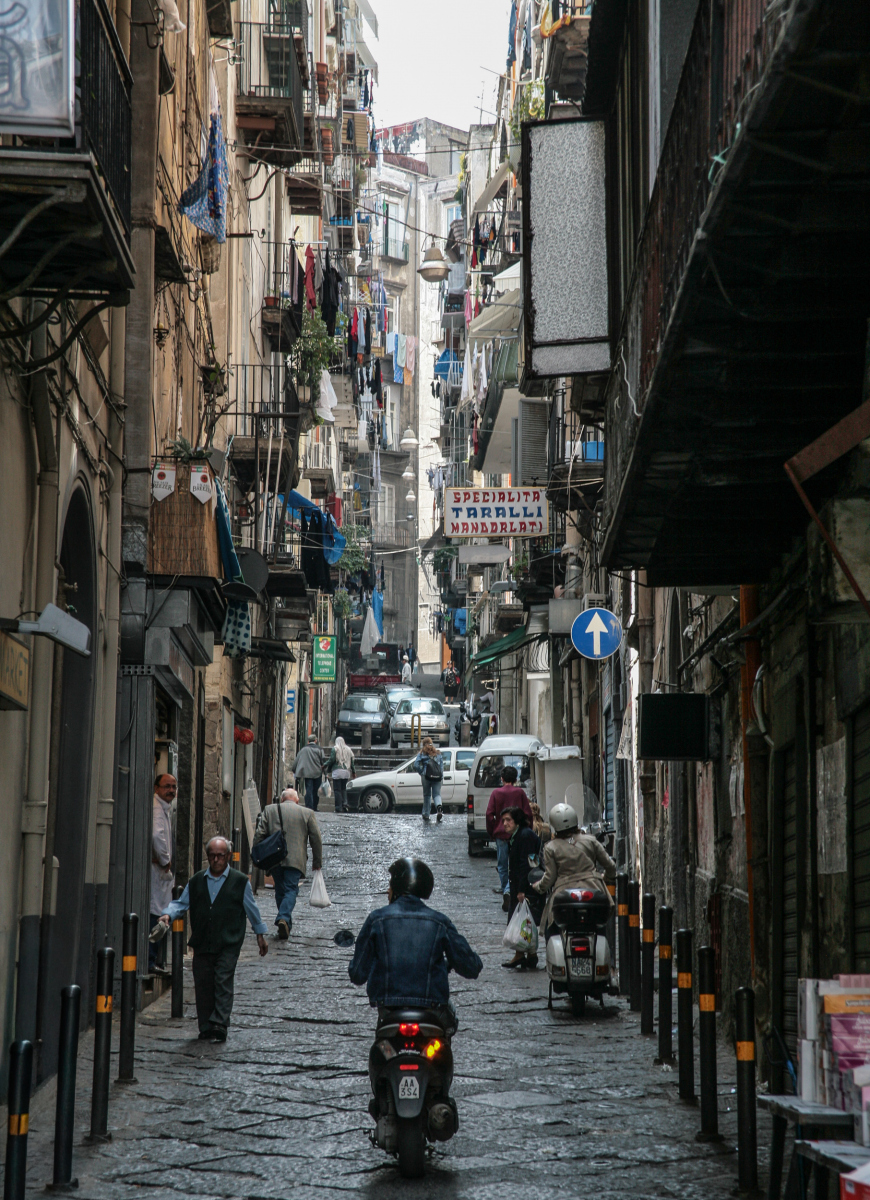 Die Gasse befindet sich im Herzen der Altstadt von Neapel, einem UNESCO-Weltkulturerbe. Solche Gassen sind das pulsierende Zentrum des neapolitanischen Lebens, wo sich Einheimische und Touristen mischen.Beschilderung: Ein Schild auf der rechten Seite weist auf "SPECIALITA TARALLI MANDORLATI" hin. Taralli sind traditionelles italienisches Knabbergebäck, das in Neapel beliebt ist und oft mit Mandeln (mandorlati) hergestellt wird.Atmosphäre: Die Szene zeigt das typische Chaos und die Energie Neapels, mit Fußgängern, Mopeds und dem Alltagsleben, das sich auf die Straße erstreckt.