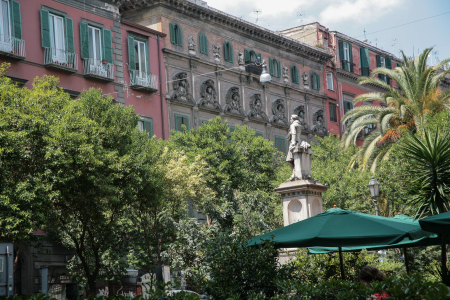 Piazza Bellini in Neapel, Italien.Lage: Der Platz liegt im historischen Zentrum von Neapel, in der Nähe der Via dei Tribunali und der Via San Sebastiano.Atmosphäre: Er ist bekannt für seine lebhafte Atmosphäre mit zahlreichen Cafés, Restaurants (wie das auf einem Schild im Vordergrund teilweise sichtbare) und der Statue in der Mitte, die den Komponisten Vincenzo Bellini darstellt.Archäologie: Eine Besonderheit des Platzes sind die freigelegten Überreste der antiken griechischen Stadtmauern, die im Boden zu sehen sind.