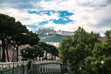 Das Gebäude auf dem Hügel im Hintergrund ist das Castel Sant'Elmo, eine mittelalterliche Festung. Die sternförmige Festung steht auf dem Vomero-Hügel, einem der höchsten Punkte der Stadt. Sie ist vor allem für ihren atemberaubenden Panoramablick über die Stadt, den Golf von Neapel und den Vesuv bekannt